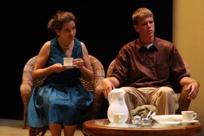 Two student actors in costume sit in armchairs on the Umble Center stage and drink tea