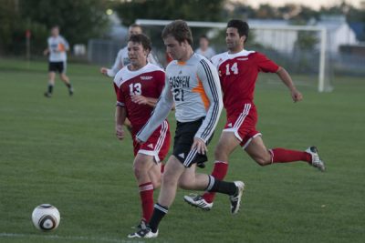 Jan Dohnal wears a white Goshen jersey and the number "21" as he kicks a soccer ball away from two players in red jerseys