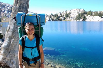 Lydia Alderfer wears a backpacking pack and poses for a picture in front of a lake in Oregon