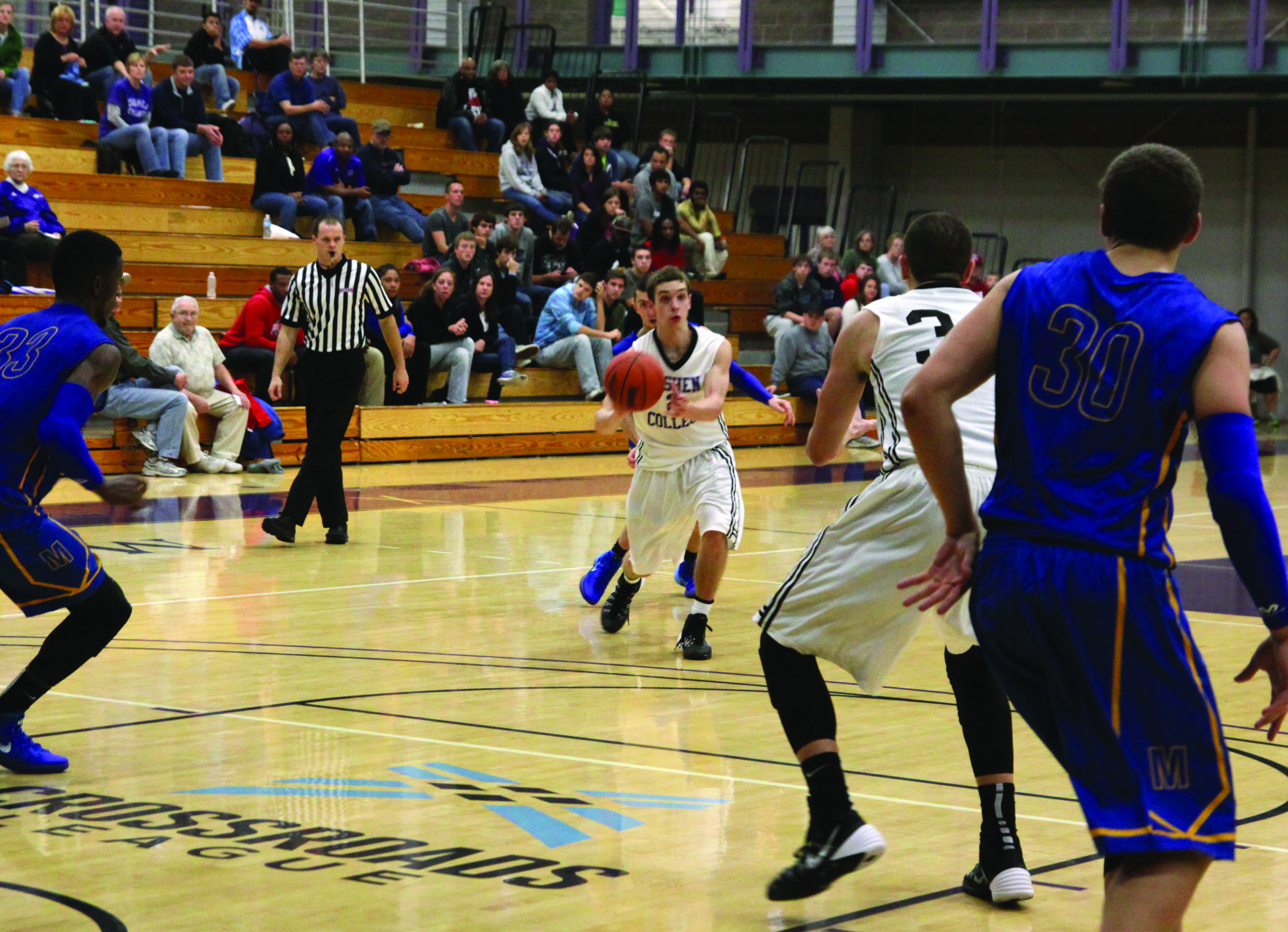 A player on the Goshen men's basketball team prepares to throw the ball in a game. Another Goshen player and two opposing players are in the foreground of the shot, while spectators can be seen on bleachers in the background