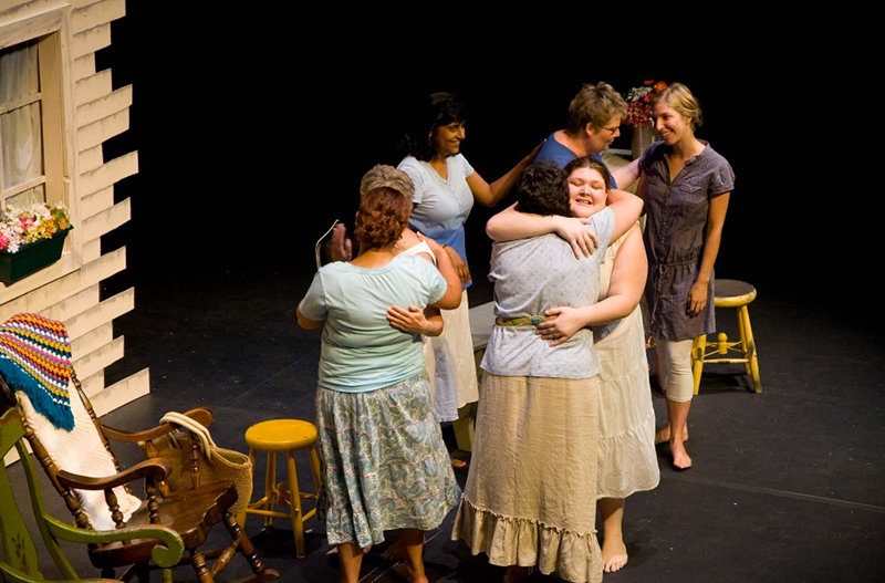 Seven female actors perform onstage in the play "Heavenly Voices." Some of the women embrace and laugh.