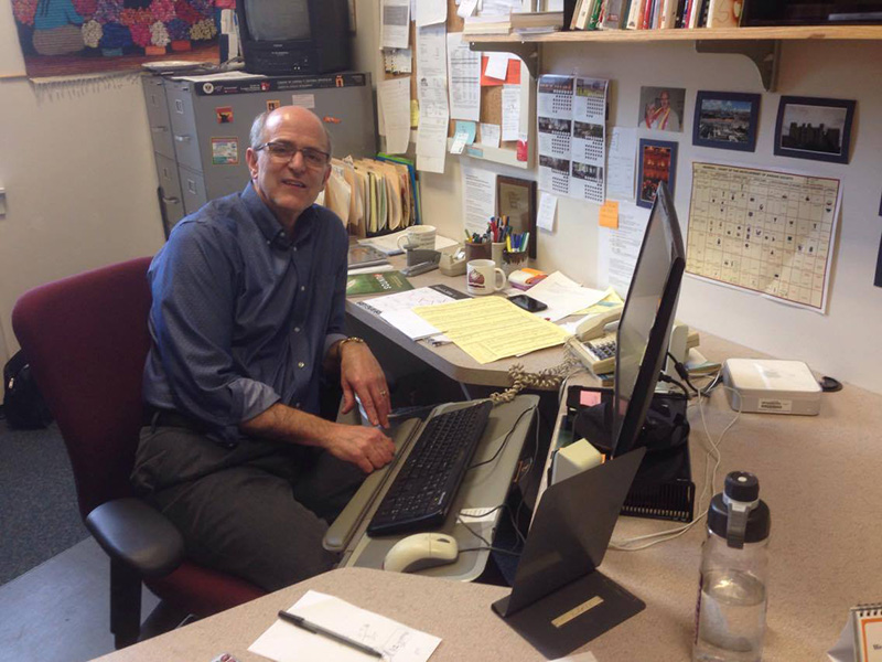 Dean Rhodes sits at a cluttered desk in his office. The walls and desk are covered in pictures, calendars, and other papers