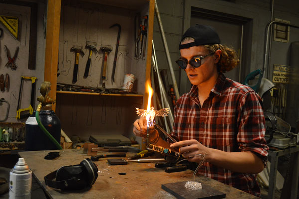 David Jantz holds one of his glass structures in a flame. He is wearing safety goggles, a backwards baseball cap, and a flannel. He is sitting in a workshop with many different tools hanging on the walls