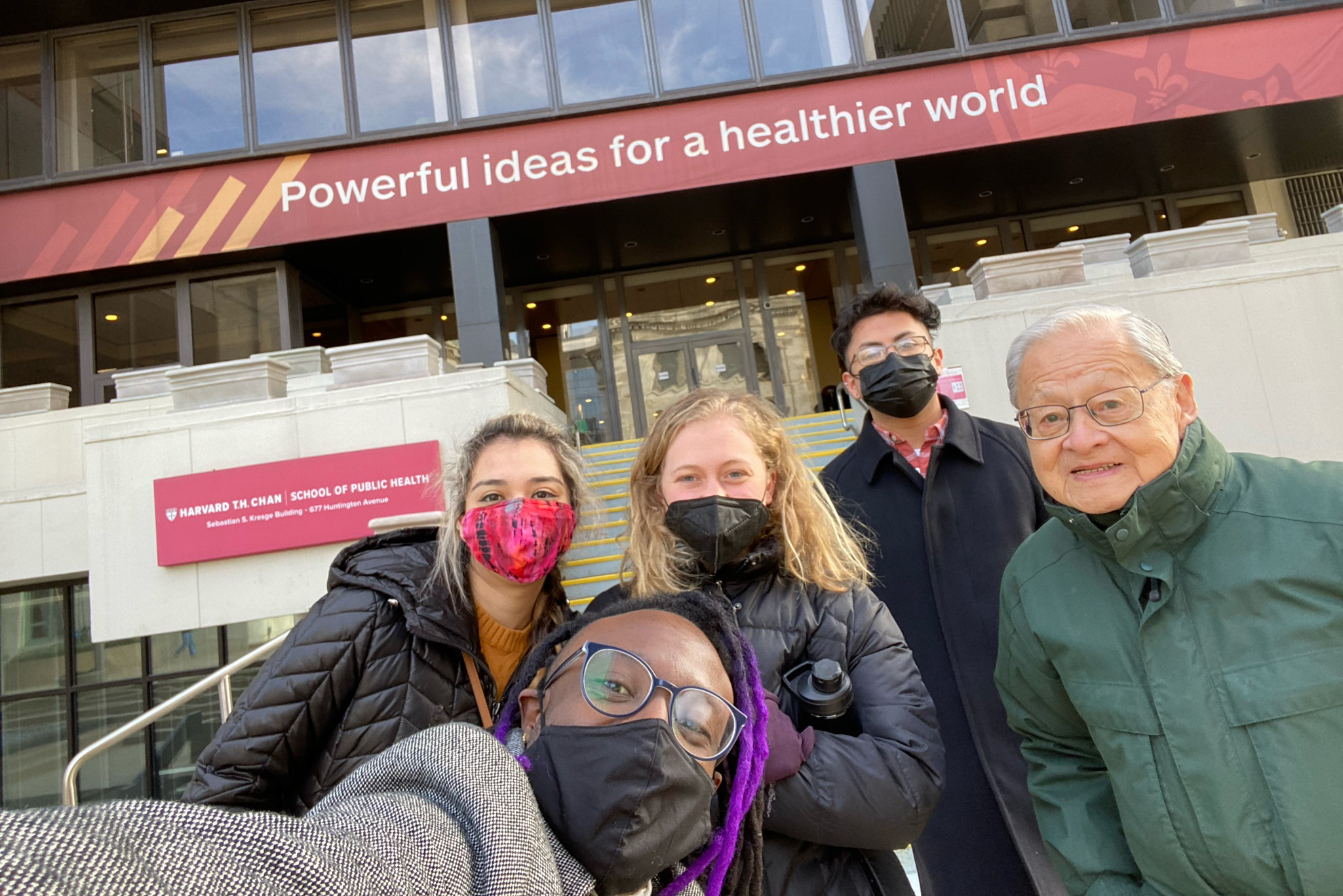 Students pose for a selfie under a Harvard building reading: "Powerful ideas for a healthier world."