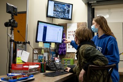 A student points to something on a computer screen while another student watches.