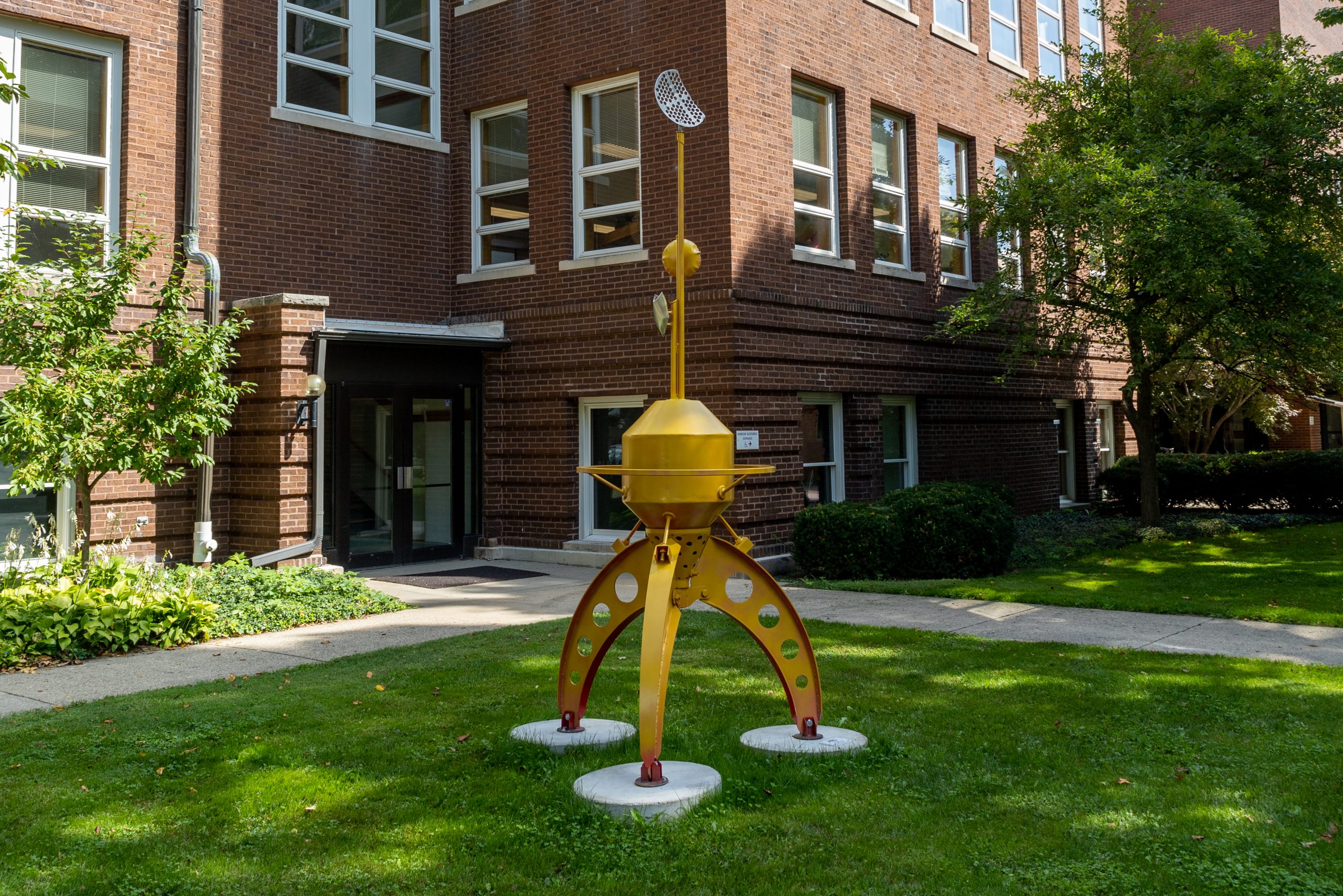A yellow scuplture sits on the lawn in front of the science building. The yellow sculpture features several legs supporting a mid section with a few long pieces coming off the top.