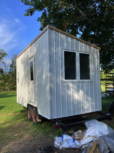 A small 18 foot trailer holds a small structure. On the side above the wheels we see a door. On the near side is a set of double windows. The roof slants up to the right. 