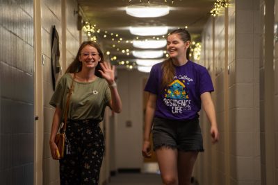 Two students walk down a dorm hallway towards the camera. one of them is waving, both are smiling.